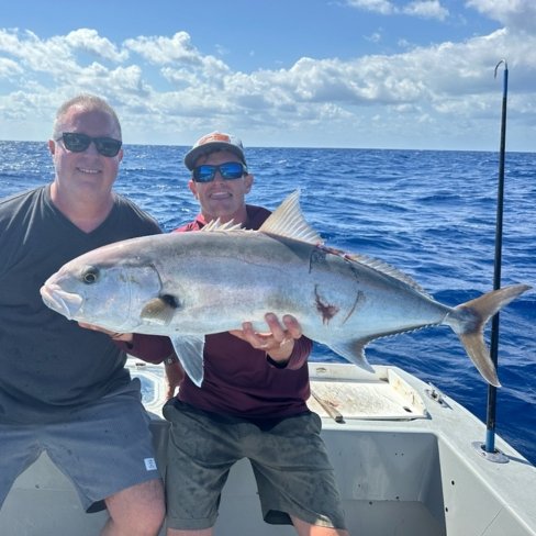 Two anglers holding a large amberjack on a fishing charter boat under a clear blue sky.
