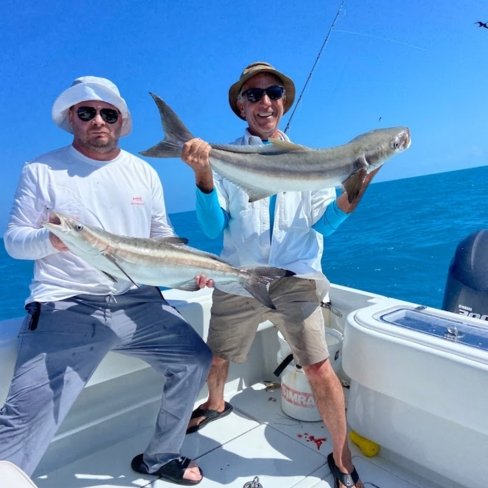 Two anglers holding large cobia fish aboard a fishing charter boat on clear blue waters.