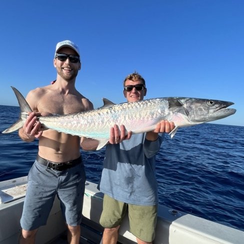 Two anglers holding a large kingfish aboard a fishing charter boat under a bright blue sky.
