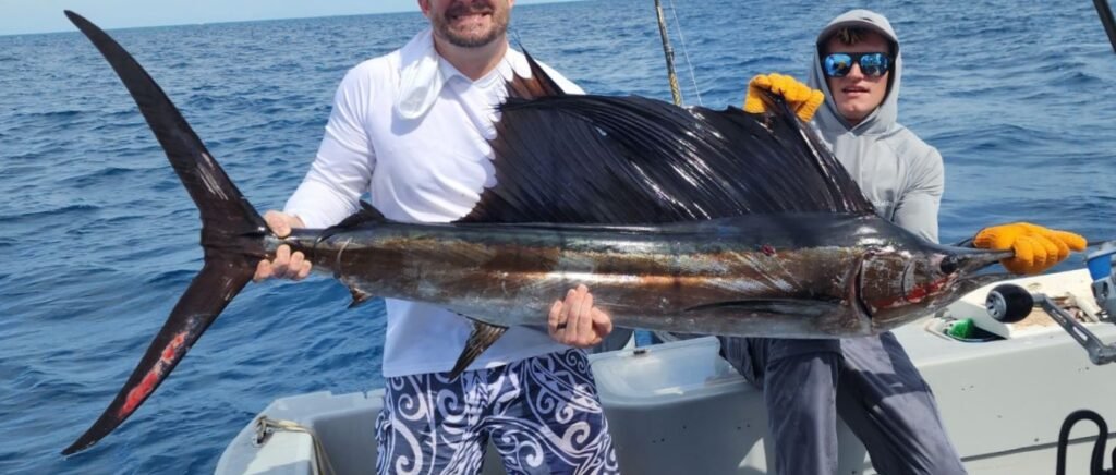 Two anglers proudly holding a large sailfish with its distinctive dorsal fin on a boat in the open ocean.